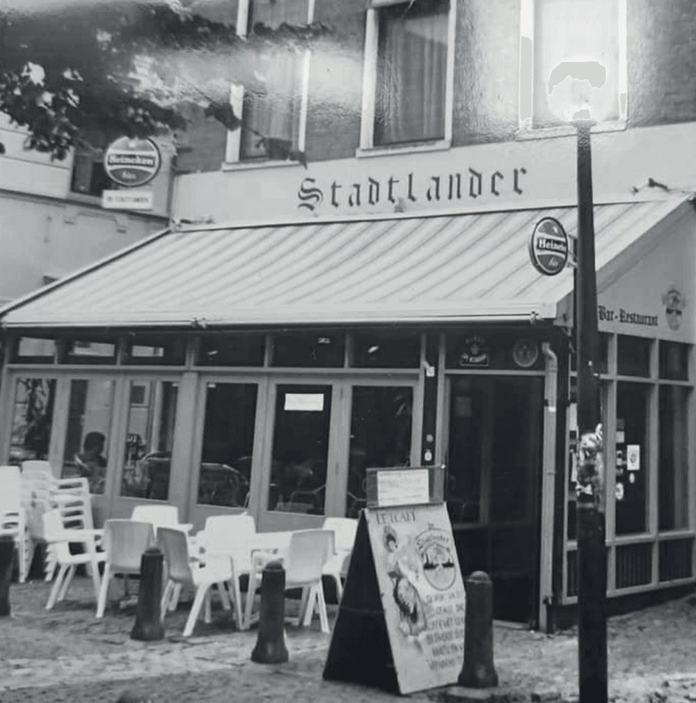 Zwart-witfoto van de oorspronkelijke pui van Stadtlander aan de Poelestraat, met gotische belettering, luifel en terrasstoelen.